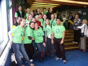 A group of students wearing green shirts gather around Jane Goodall, a famous anthropologist, for a photo.