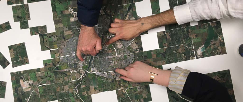 An aerial map has been cut into pieces to create a puzzle. Some pieces of the puzzle have been put back together, others remain disjointed. There are three light skinned hands in the frame. One is older than the others, another has a tattoo on its wrist, the third is wearing a bracelet. The hands point to different areas of the map, as if in conversation with each other.