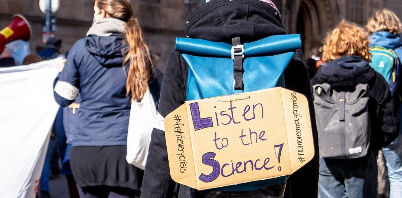 A cardboard sign with the words "Listen to the Science" written on it is taped to the back of a woman's backpack, as she walks through a crowd